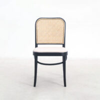 A wooden chair with a black frame, a woven cane backrest, and a light-coloured cushioned seat, positioned on a plain light floor against a white background.