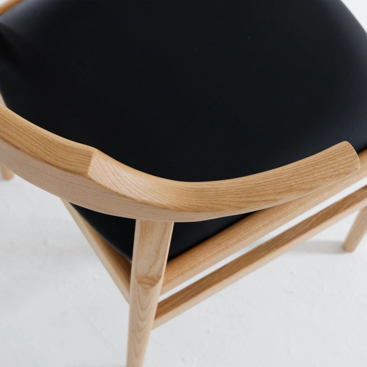 Final Chair_B4 A close-up of a wooden chair with a light wood frame and a black cushioned seat, set against a white background. The image highlights the smooth, curved armrest and joinery of the chair.