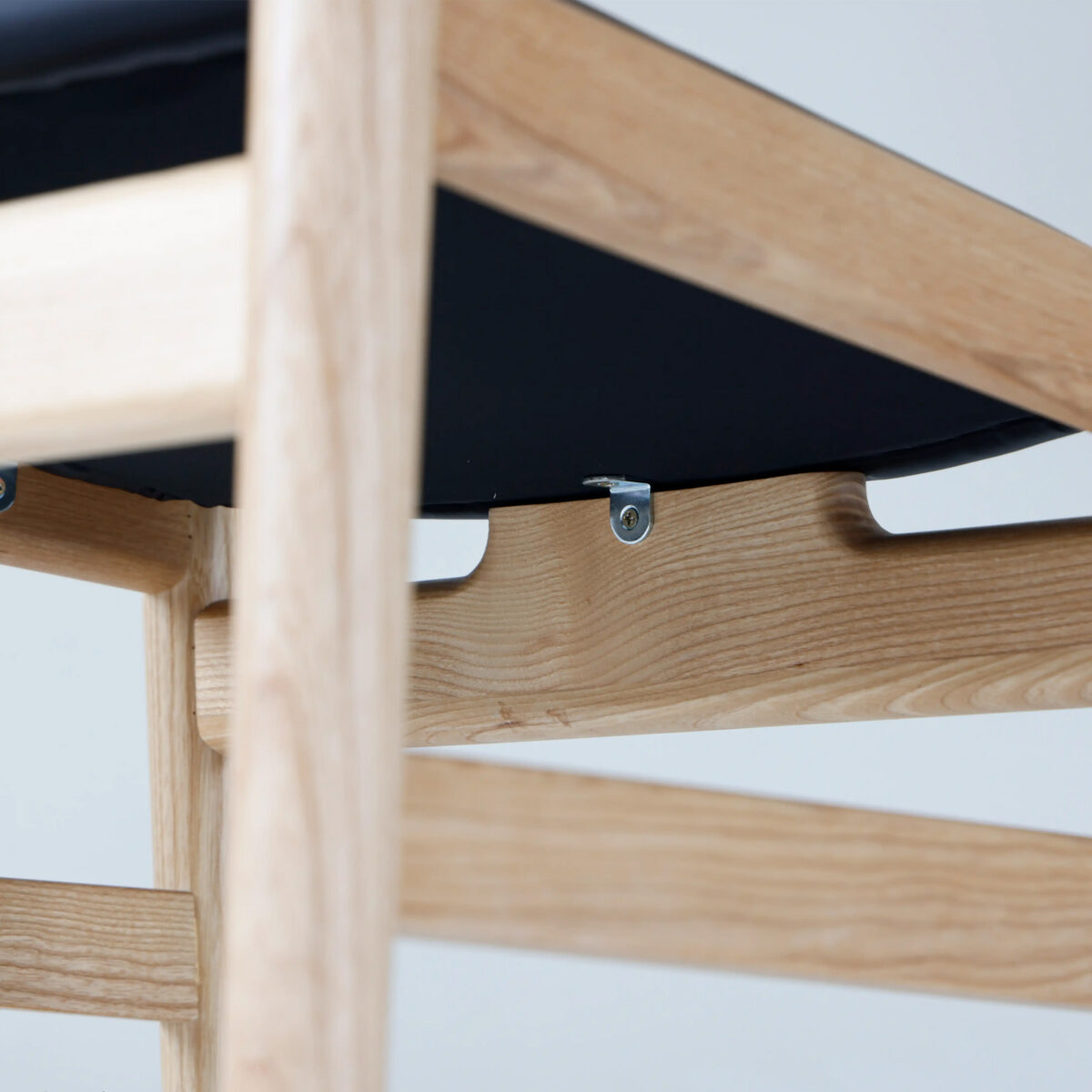 Final Chair_B6 Close-up view of the underside of a wooden chair, showing light-coloured wooden frame, metal bolts, and a black seat. The construction details and wood grain are clearly visible.