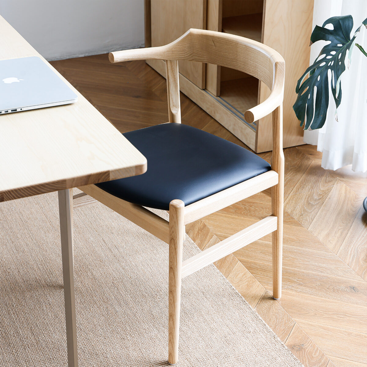 Final Chair_C3 A light wooden chair with a curved backrest and black seat cushion is placed next to a light wood desk with a laptop on it. The setting features a herringbone floor, a beige rug, and a green plant near a window.