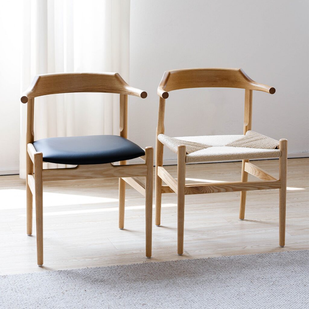 Two wooden chairs with curved backs are side by side in a sunlit room. One has a black cushioned seat, and the other has a woven seat. Light wood flooring and a white curtain are visible in the background.