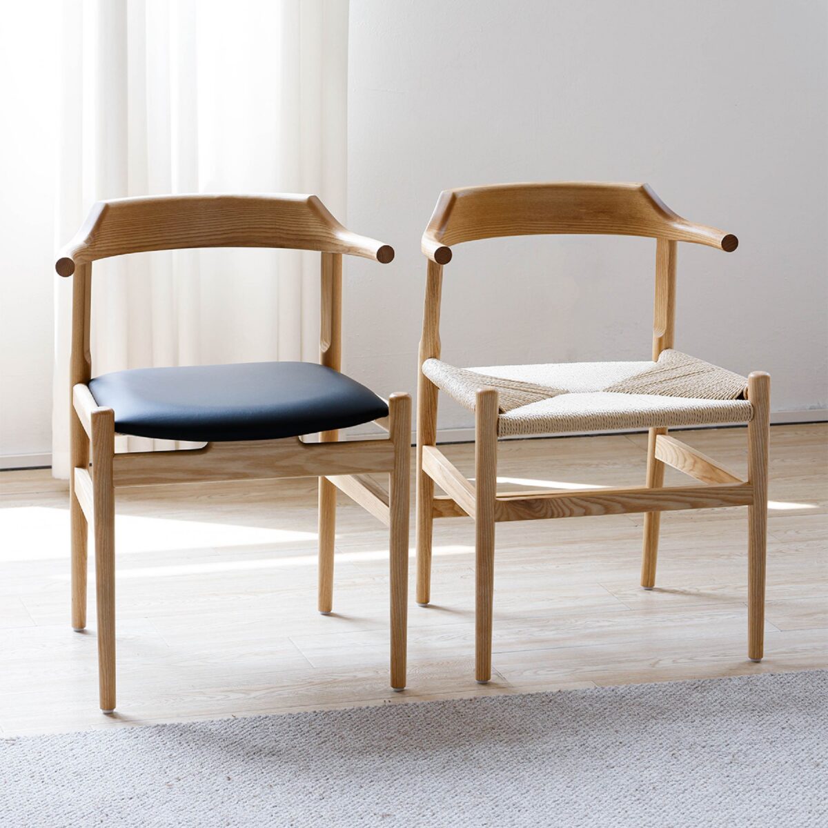 Final Chair_C5 Two wooden chairs with curved backs are side by side in a sunlit room. One has a black cushioned seat, and the other has a woven seat. Light wood flooring and a white curtain are visible in the background.