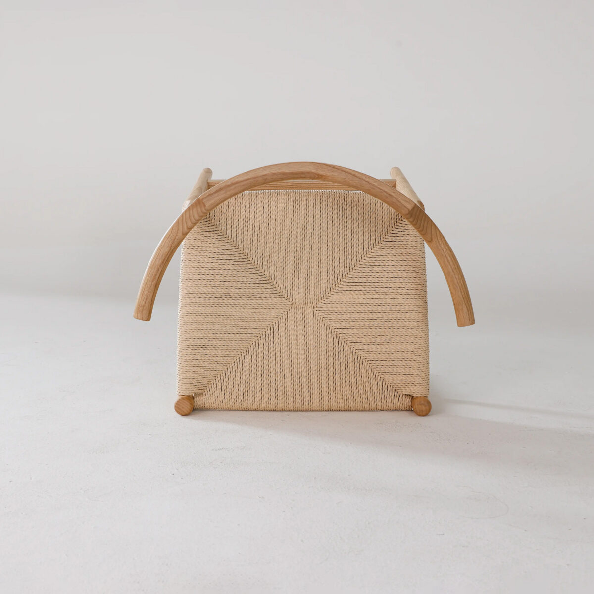 Final Chair_Paper Robe_B10 A woven chair is shown upside down on a light surface, revealing its square, textured seat, wooden frame, and curved wooden armrests. The background is plain and neutral.