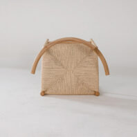A woven chair is shown upside down on a light surface, revealing its square, textured seat, wooden frame, and curved wooden armrests. The background is plain and neutral.