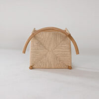 A woven chair is shown upside down on a light surface, revealing its square, textured seat, wooden frame, and curved wooden armrests. The background is plain and neutral.