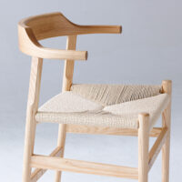 A light wooden chair with curved armrests and a woven seat, displayed against a plain, light grey background. The design is modern and minimalistic.
