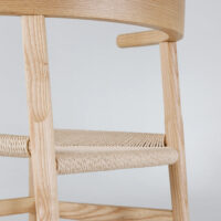 Close-up of a wooden chair with a woven seat and rounded backrest, showcasing light-coloured wood grain and smooth craftsmanship against a plain, light background.