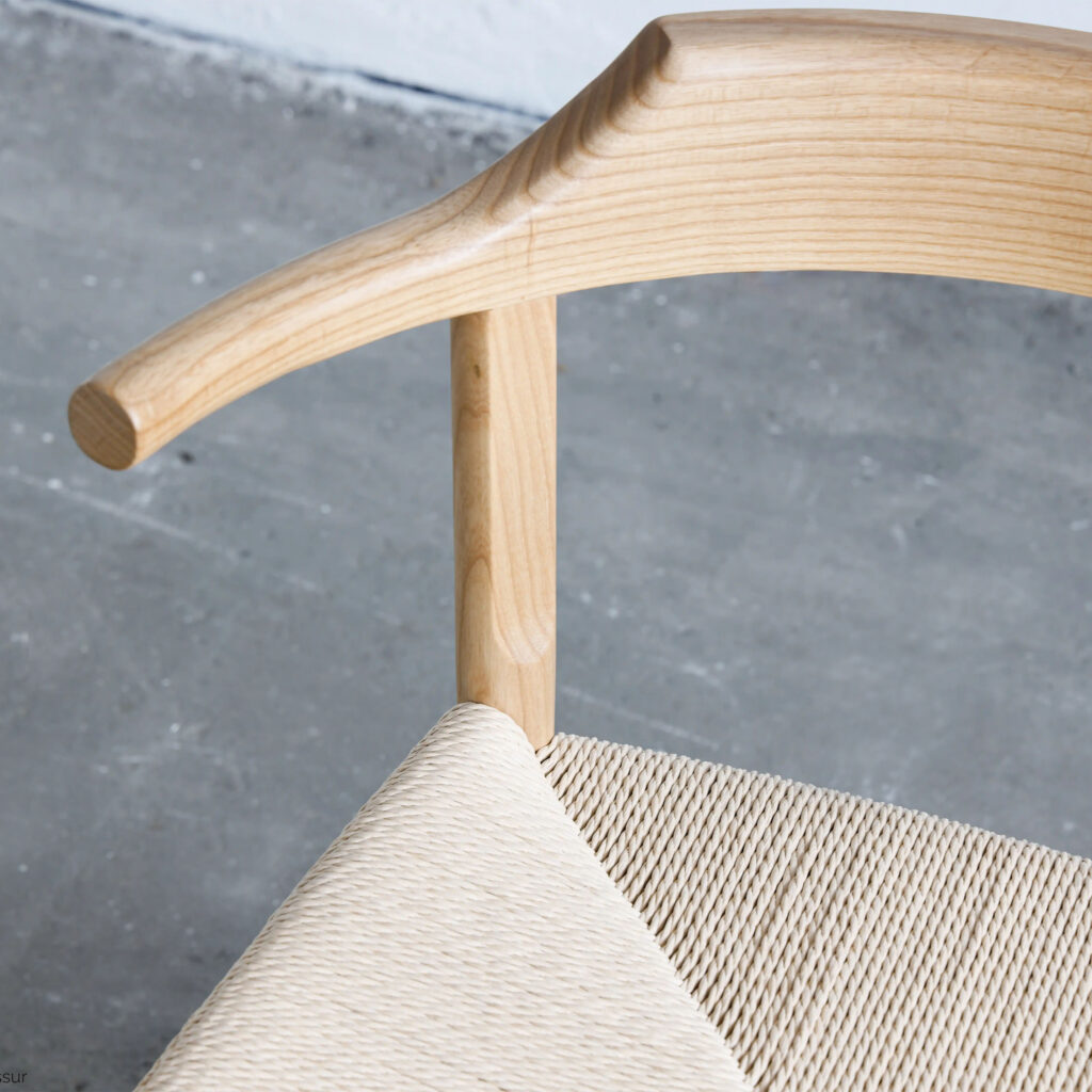 Close-up of a Scandinavian-style wooden chair with a curved backrest and woven seat, set on a grey concrete floor. The wood has a smooth light finish, highlighting the chair's minimalist design.