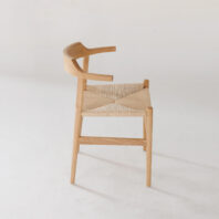 A minimalist wooden chair with a curved backrest and woven seat, placed on a plain white background. The chair features clean lines and a natural finish, highlighting its modern design.