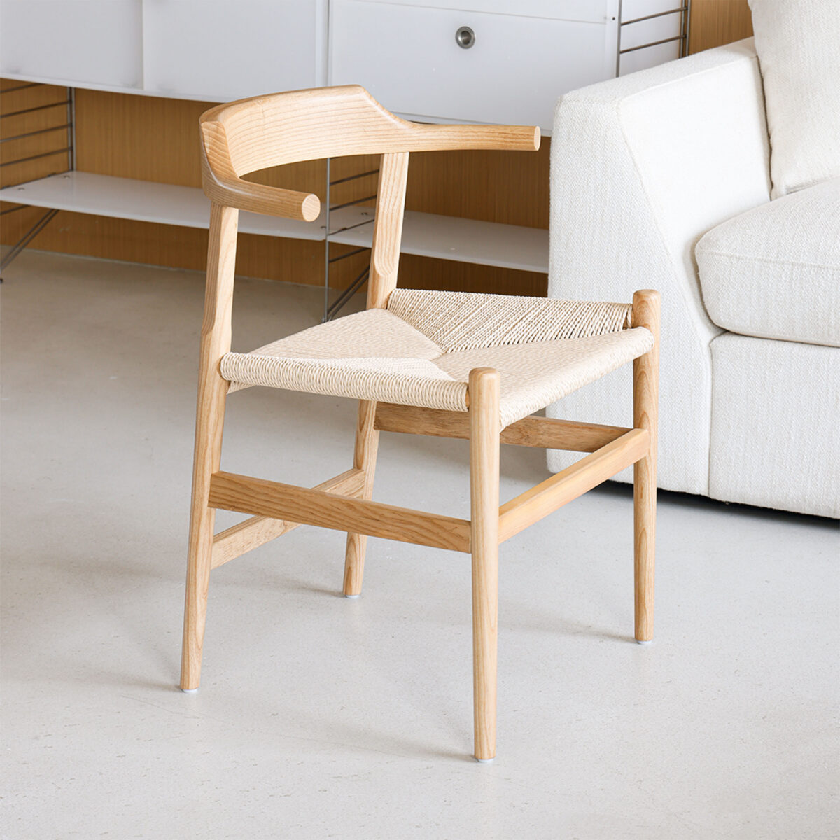 Final Chair_Paper Robe_C1 A modern wooden chair with a woven seat and curved backrest, placed on a light-coloured floor next to a white sofa and a white cupboard.