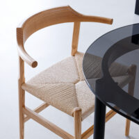 A wooden chair with a curved backrest and woven seat is positioned next to a round black glass table on a light-coloured floor.