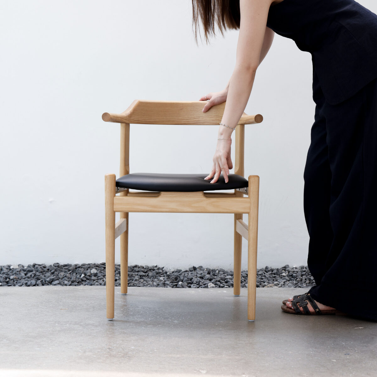 Final_Outdoor26 A person in black clothing stands next to a wooden chair with a black seat, placing one hand on the chair’s backrest and another on the seat, against a white wall background with stones on the ground.
