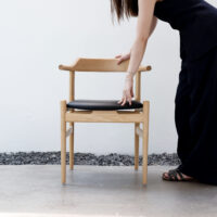 A person in black clothing stands next to a wooden chair with a black seat, placing one hand on the chair’s backrest and another on the seat, against a white wall background with stones on the ground.