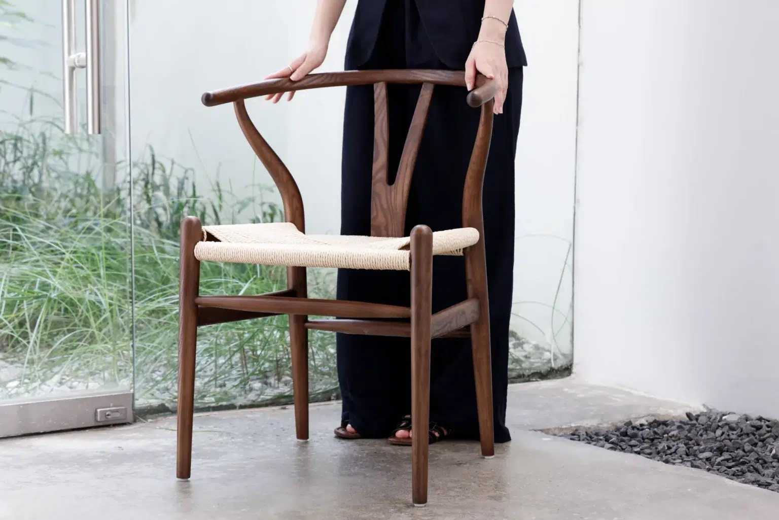 Outdoor10 A person in dark clothing stands indoors, holding a wooden chair with a woven seat. The background features a glass wall with plants visible outside.