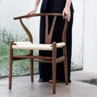 A person in dark clothing stands indoors, holding a wooden chair with a woven seat. The background features a glass wall with plants visible outside.