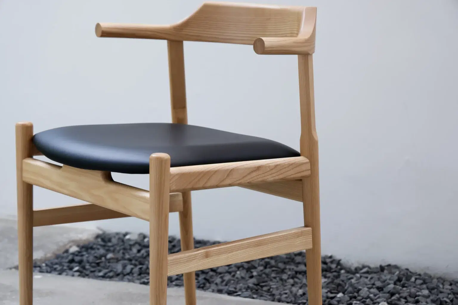 Outdoor25 A modern wooden chair with a light finish, black cushioned seat, and minimalist armrests is placed on a concrete floor next to a bed of dark pebbles against a plain white wall.