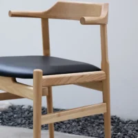 A modern wooden chair with a light finish, black cushioned seat, and minimalist armrests is placed on a concrete floor next to a bed of dark pebbles against a plain white wall.