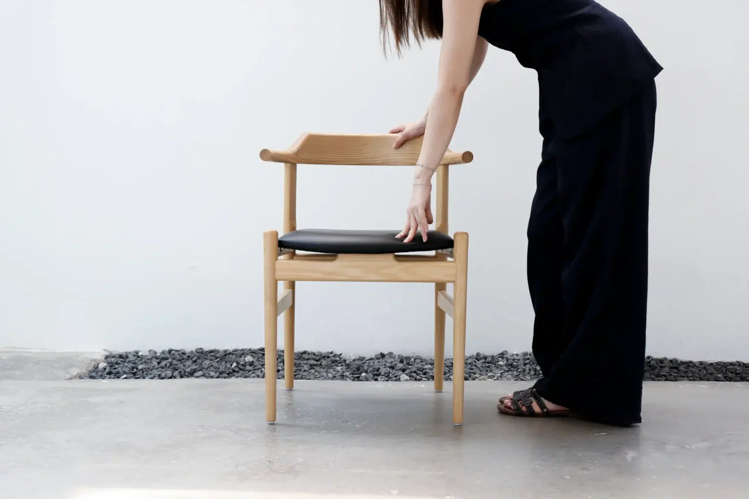 Outdoor26 A woman dressed in black leans forward to place or adjust a wooden chair with a black cushion on a concrete floor, against a white wall and a strip of small black stones.