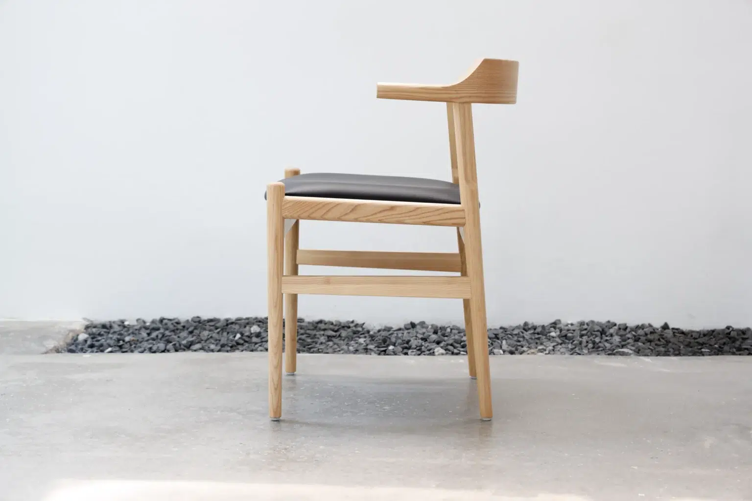 Outdoor27 A modern wooden chair with a light wood frame, black cushioned seat, and one raised armrest, set on a polished concrete floor beside a line of dark stones against a white wall.