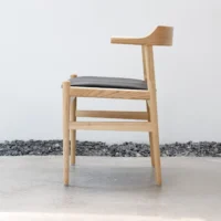 A modern wooden chair with a light wood frame, black cushioned seat, and one raised armrest, set on a polished concrete floor beside a line of dark stones against a white wall.