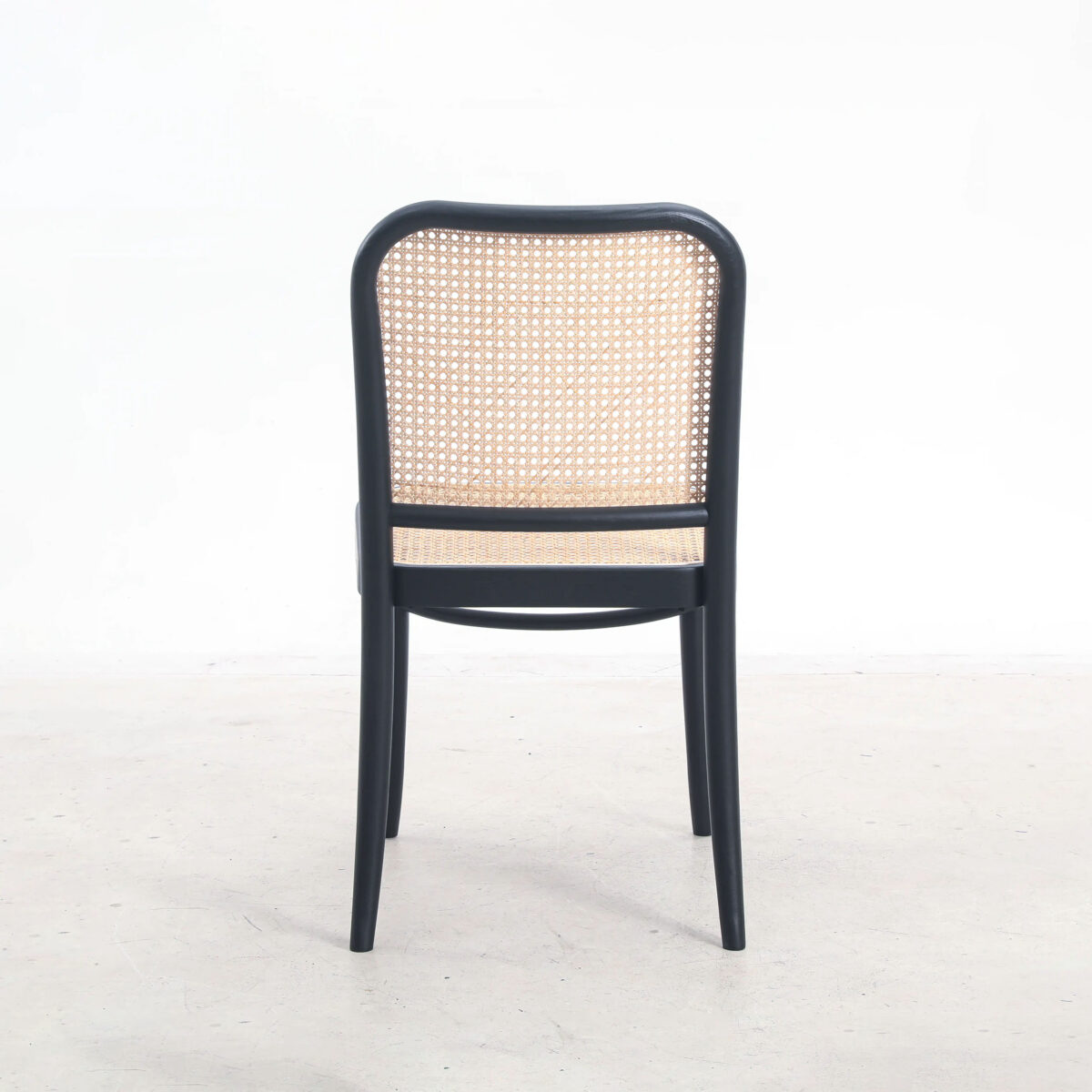 Rattan_B2 A wooden chair with a black frame and a woven cane backrest, viewed from behind, is placed on a light-coloured floor against a plain white background.
