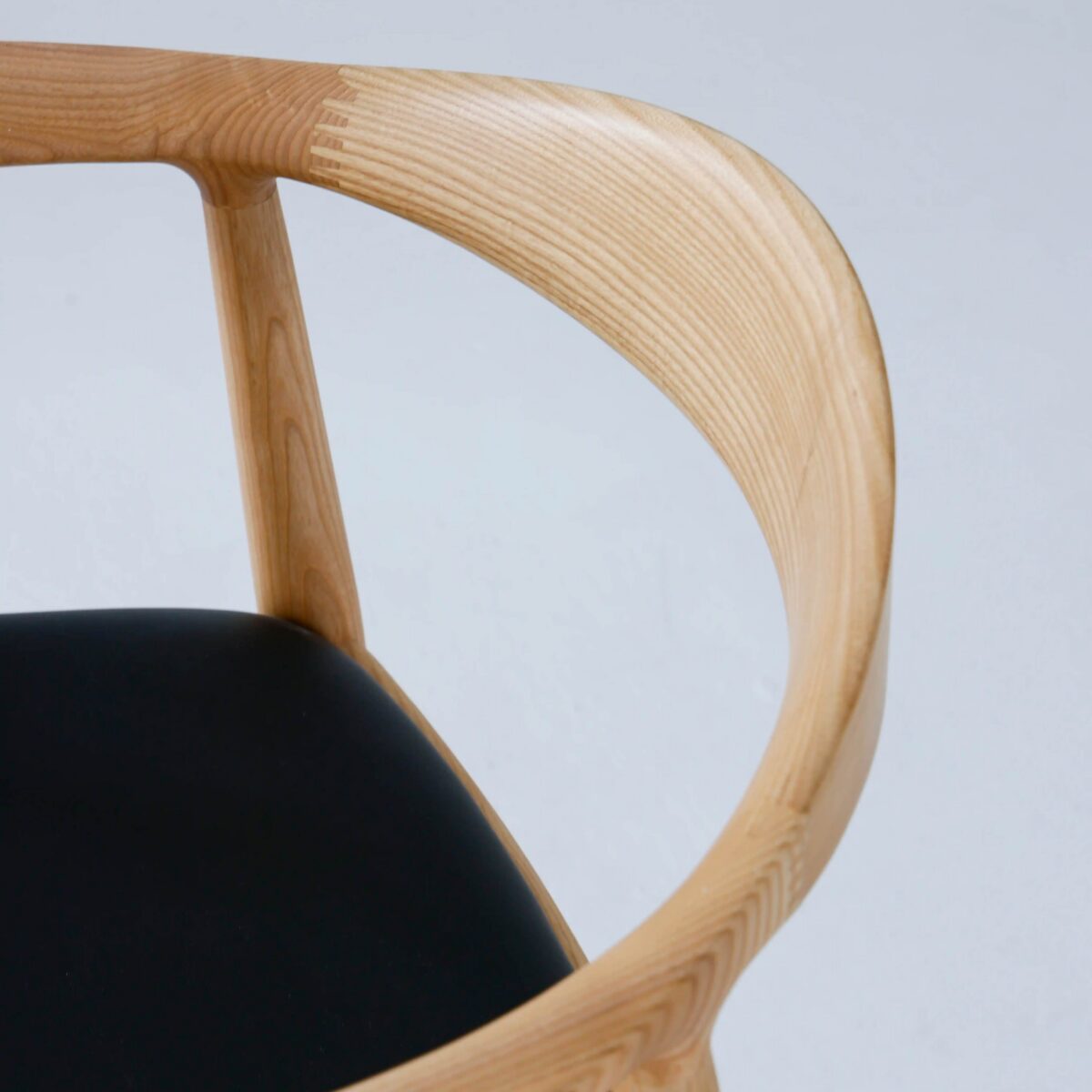 Round Chair_Leather_B10 A close-up view of a wooden chair with a curved, smooth armrest and a black cushioned seat, set against a plain light background.