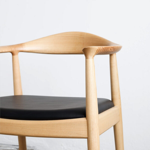A close-up of a modern wooden chair with a curved backrest and black cushioned seat, set against a plain light-coloured background.