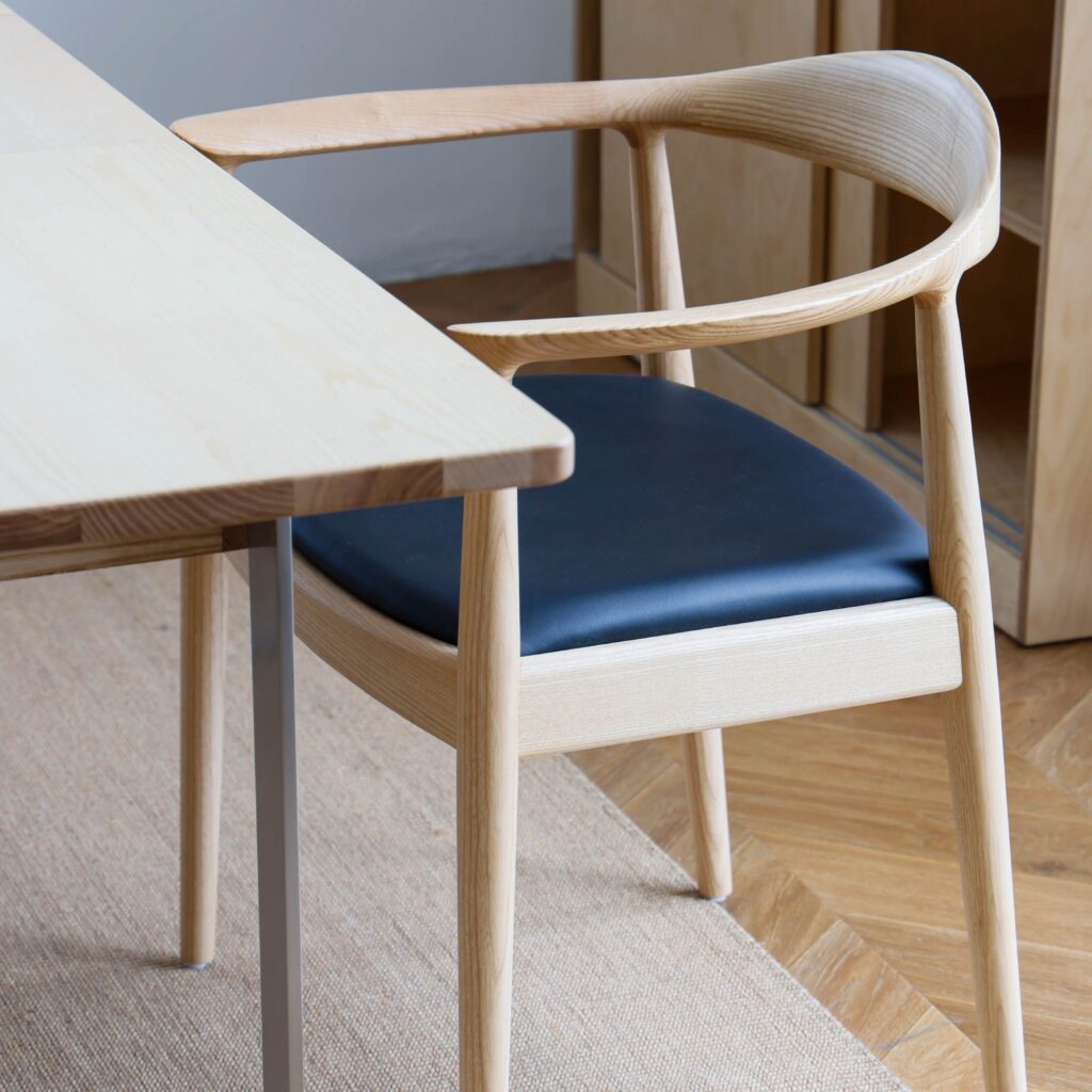 A light wood chair with curved armrests and a black cushioned seat is placed next to a light wood table on a beige rug, in a room with wooden flooring.