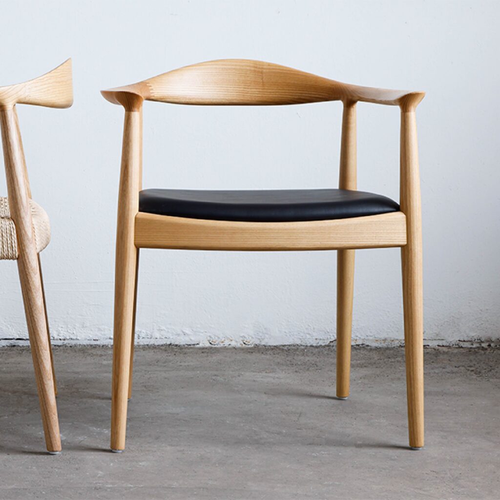 A modern wooden chair with curved armrests and a black cushioned seat sits on a concrete floor against a white wall. Part of another similar chair is visible on the left.