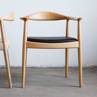 A modern wooden chair with curved armrests and a black cushioned seat sits on a concrete floor against a white wall. Part of another similar chair is visible on the left.