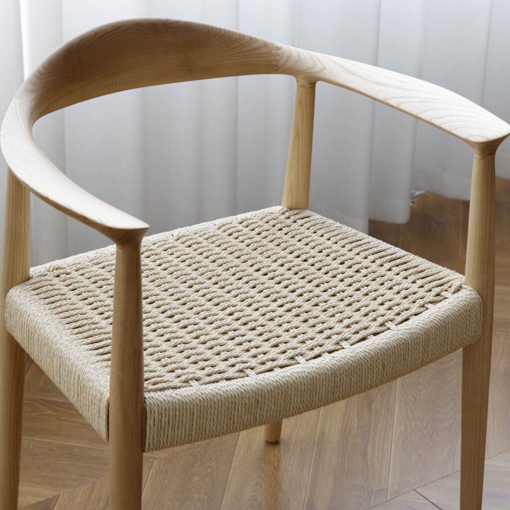 A wooden chair with a curved backrest and a woven seat, placed on a wooden floor near a window with sheer white curtains.