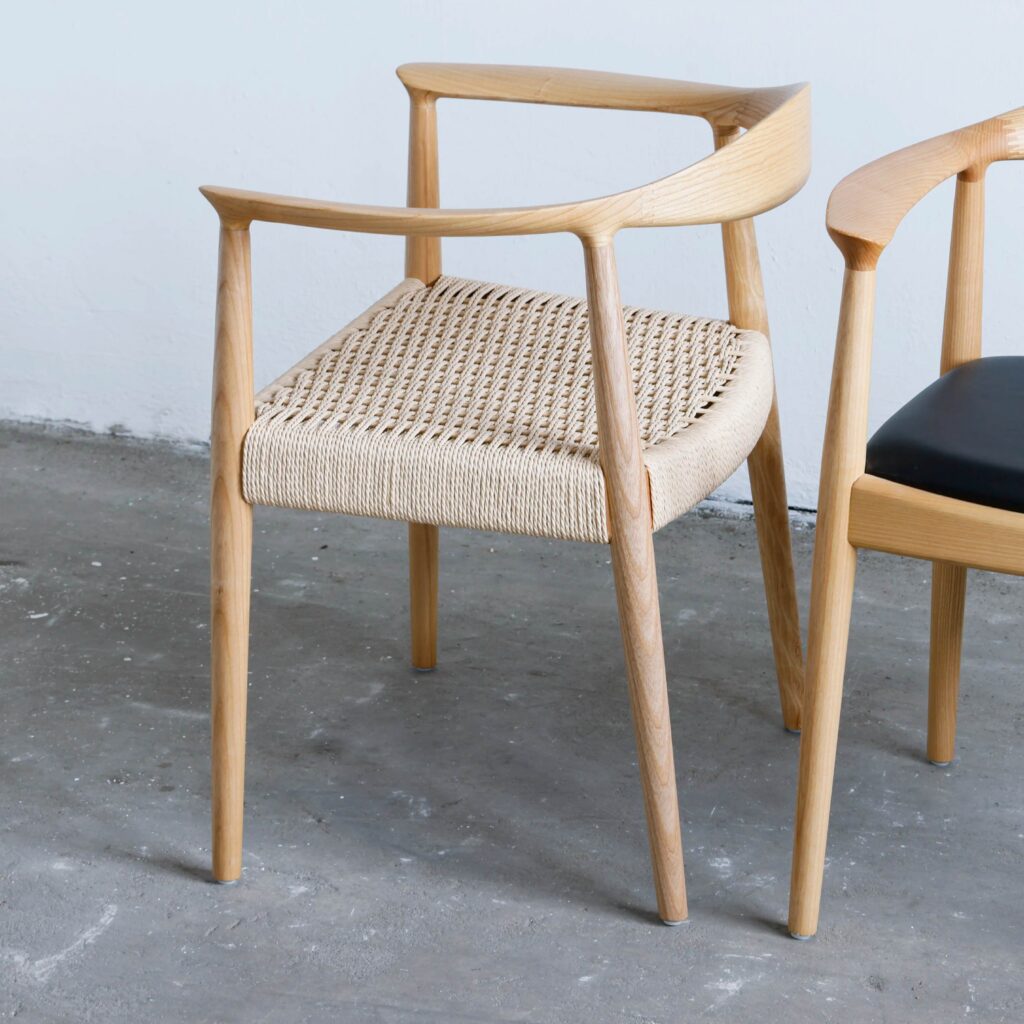 A modern wooden chair with curved armrests and a woven seat, placed on a concrete floor next to another chair with a black seat.
