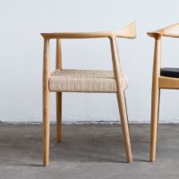 A modern wooden chair with a woven seat and curved backrest is positioned on a concrete floor next to another partially visible chair with a black seat.