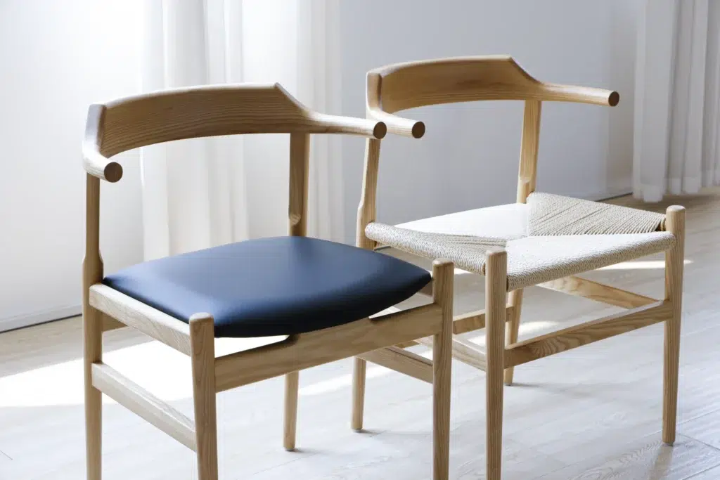 Two wooden chairs with curved backrests are side by side in a sunlit room; one chair has a black padded seat, and the other has a woven seat. Light streams through sheer curtains in the background.