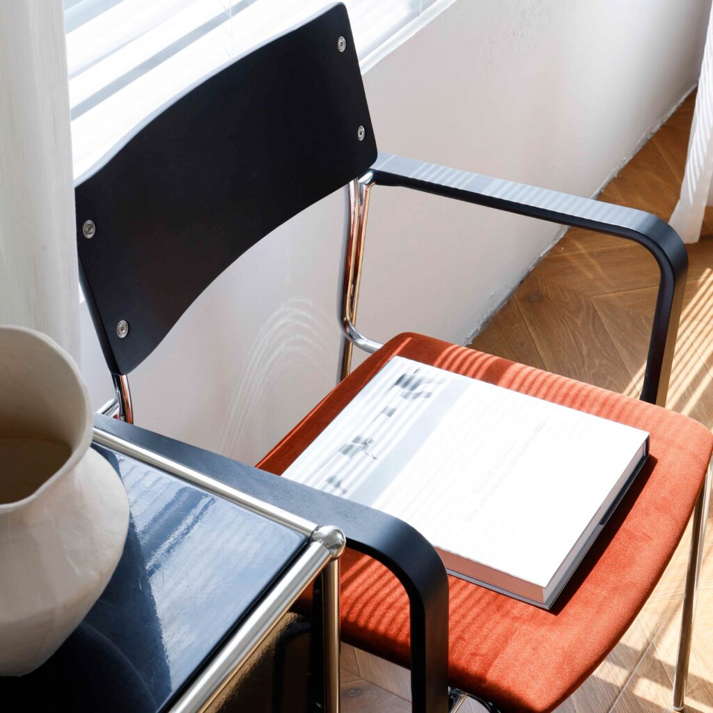 A modern chair with a black backrest and orange seat holds an open book. Next to it is a black side table with a white ceramic vase. Sunlight streams through blinds, casting shadows on the scene.