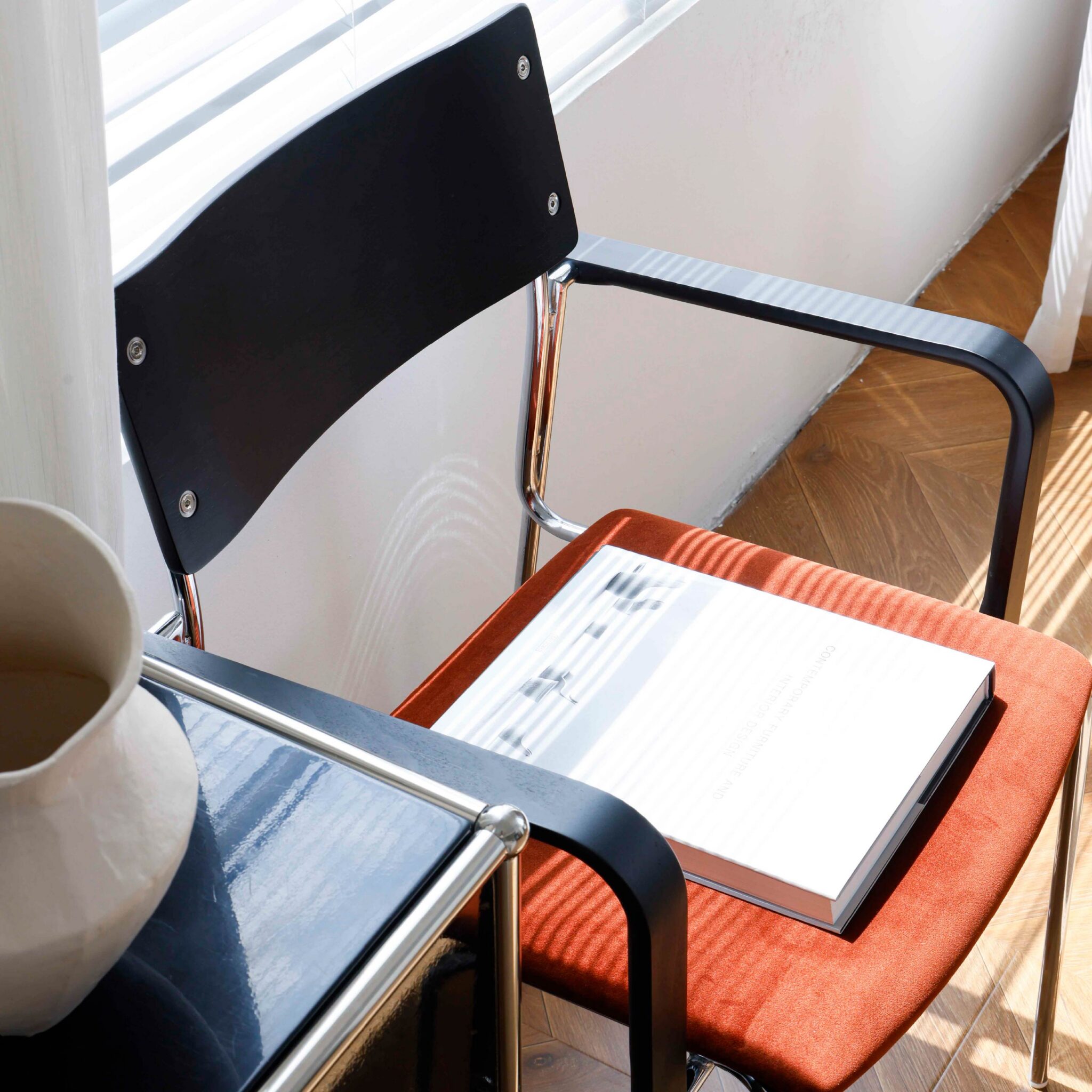 A modern chair with a black backrest and orange seat holds an open book. Next to it is a black side table with a white ceramic vase. Sunlight streams through blinds, casting shadows on the scene.