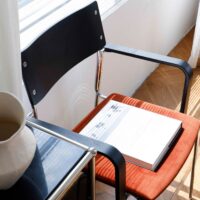 A modern chair with a black backrest and orange seat holds an open book. Next to it is a black side table with a white ceramic vase. Sunlight streams through blinds, casting shadows on the scene.