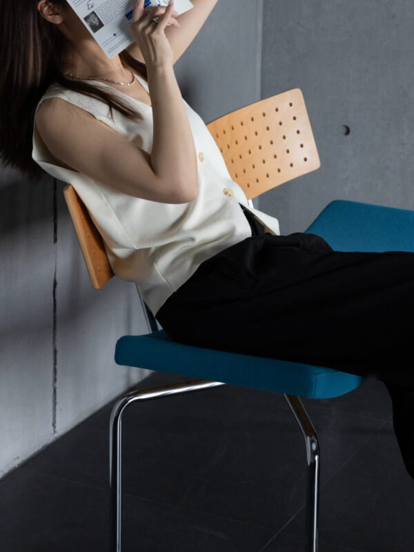 Woman sitting on modern bench seat with perforated timber backrest and blue fabric seat, holding a book over her face, concrete background.