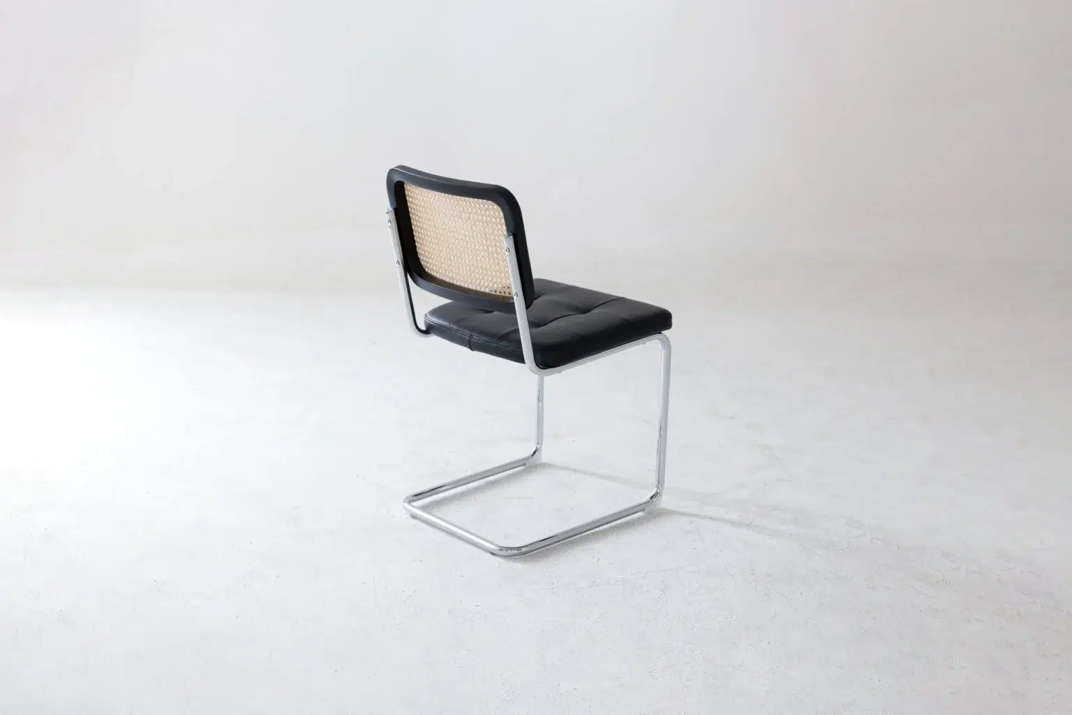 Back side view A single chair with a chrome cantilever frame, black seat cushion, and a woven cane backrest is positioned on a plain white floor against a light background.