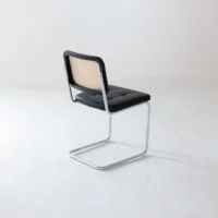 A single chair with a chrome cantilever frame, black seat cushion, and a woven cane backrest is positioned on a plain white floor against a light background.
