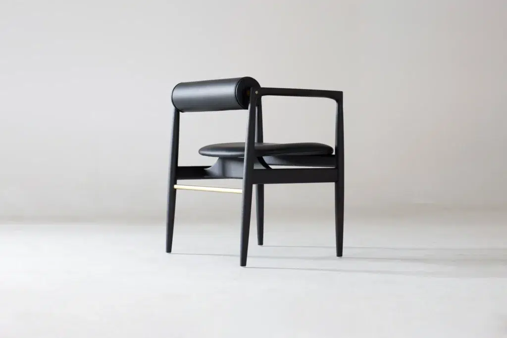A modern black chair with a cylindrical backrest, an angular frame, and a cushioned seat is placed on a light, minimalist background. The design features clean lines and a gold accent beneath the seat.