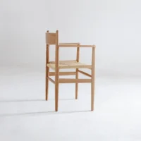 A minimalist wooden chair with a woven seat, square backrest, and simple legs, placed in a bright, empty room with a white floor and wall.