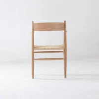 A simple wooden chair with a woven seat is positioned on a plain white floor against a white background, shown from the back.