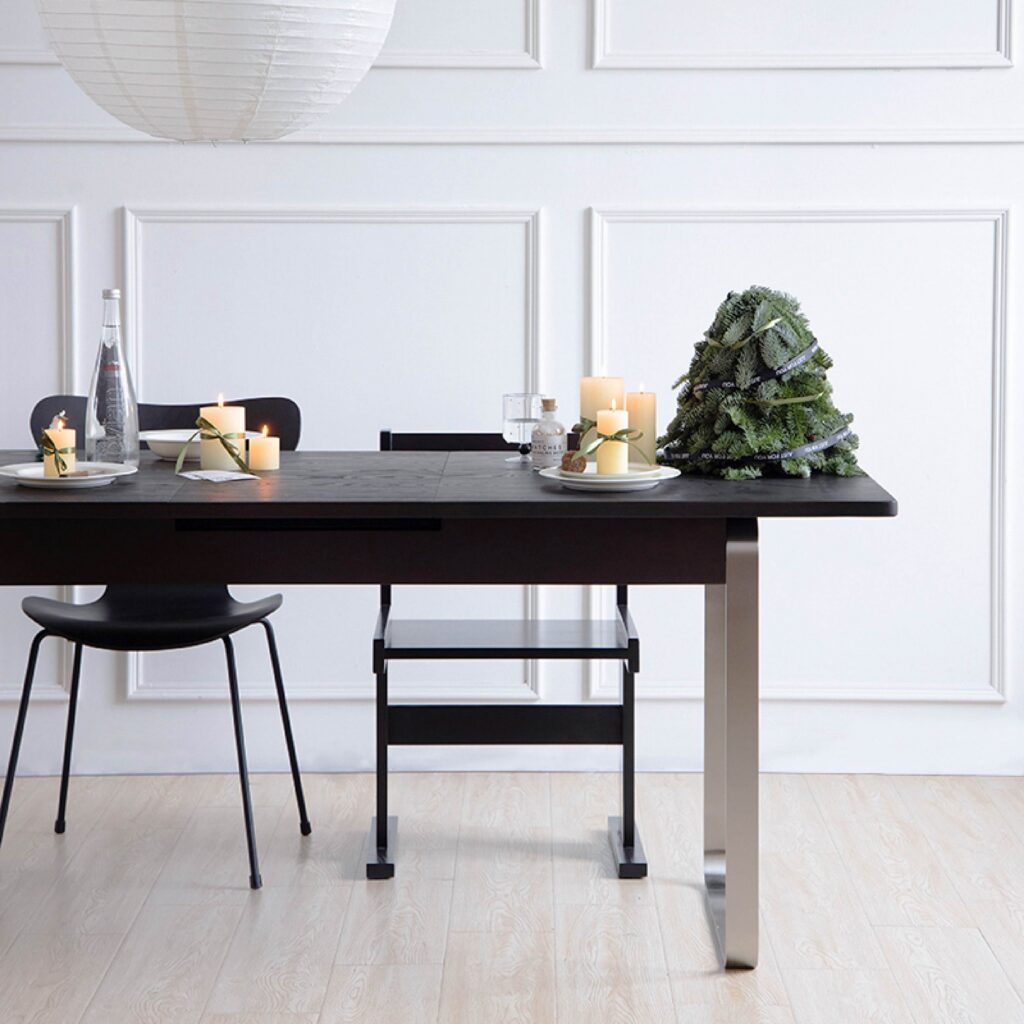 A modern dining table with black chairs, decorated with candles, wrapped presents, and a small Christmas tree centrepiece, set in a bright room with white panelled walls and light wooden flooring.