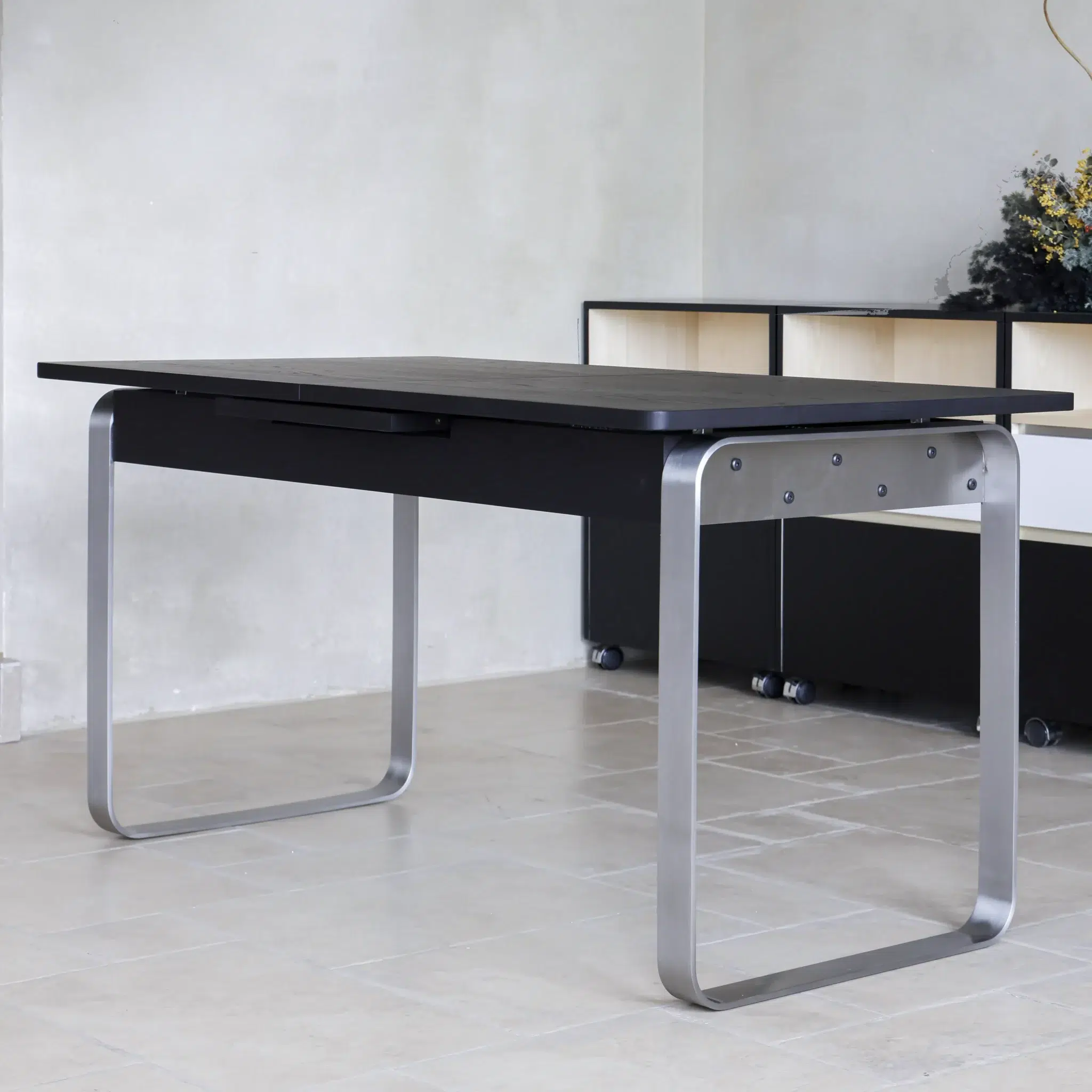 A modern desk with a black rectangular top and curved metal legs stands on a light tiled floor. In the background, there is a black and white storage unit and a vase with flowers.