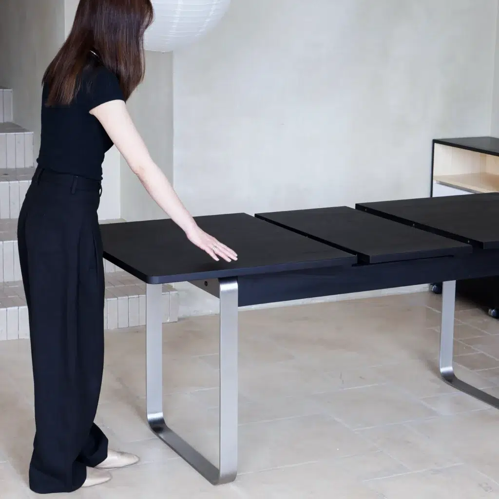 A woman in black clothing stands beside a modern black dining table with metal legs, extending its surface. The setting appears to be a minimalistic, light-coloured room with tiled floors.