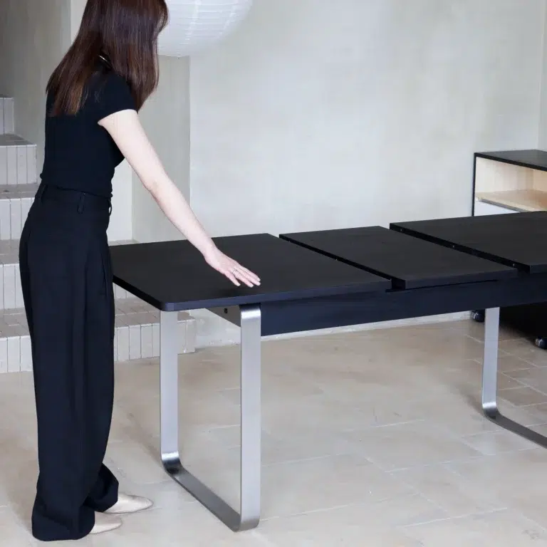 A woman in black clothing stands beside a modern black dining table with metal legs, extending its surface. The setting appears to be a minimalistic, light-coloured room with tiled floors.