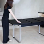 A woman in black clothing stands beside a modern black dining table with metal legs, extending its surface. The setting appears to be a minimalistic, light-coloured room with tiled floors.