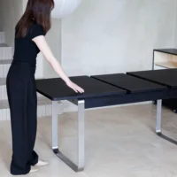 A woman in black clothing stands beside a modern black dining table with metal legs, extending its surface. The setting appears to be a minimalistic, light-coloured room with tiled floors.