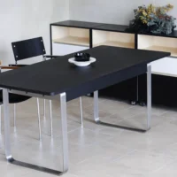 A modern black table with metal legs is set with black and white crockery. Two black chairs are on one side, and white shelves with decorative plants are in the background on a light tiled floor.